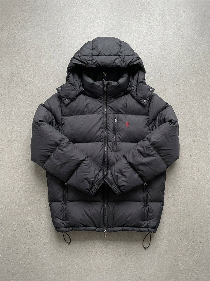The same black puffer re-shot as a flat-lay on a pale grey concrete surface with the sleeves folded inwards