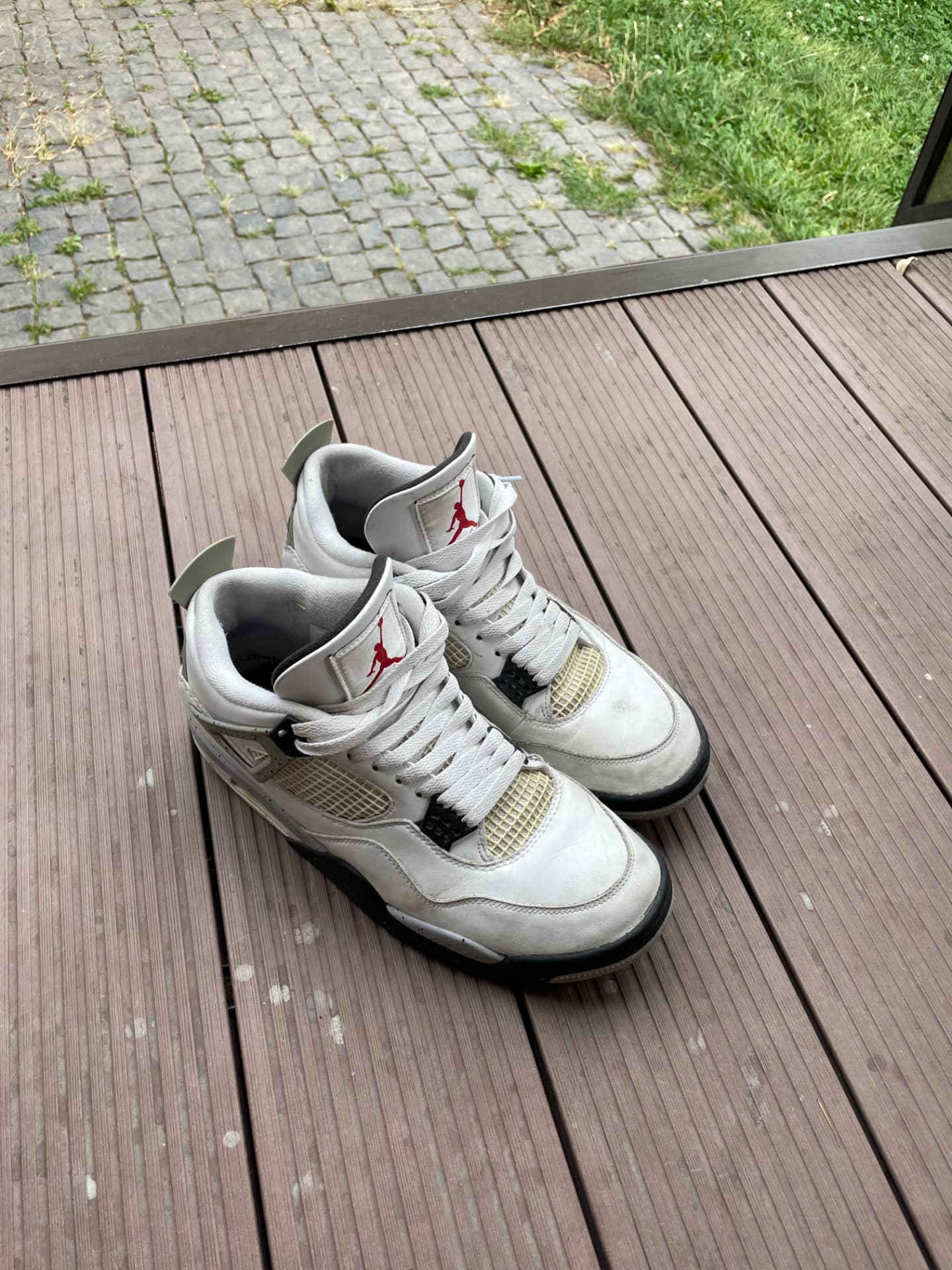 The original phone photo: a pair of white Air Jordan 4 sneakers on brown decking with paving and grass behind