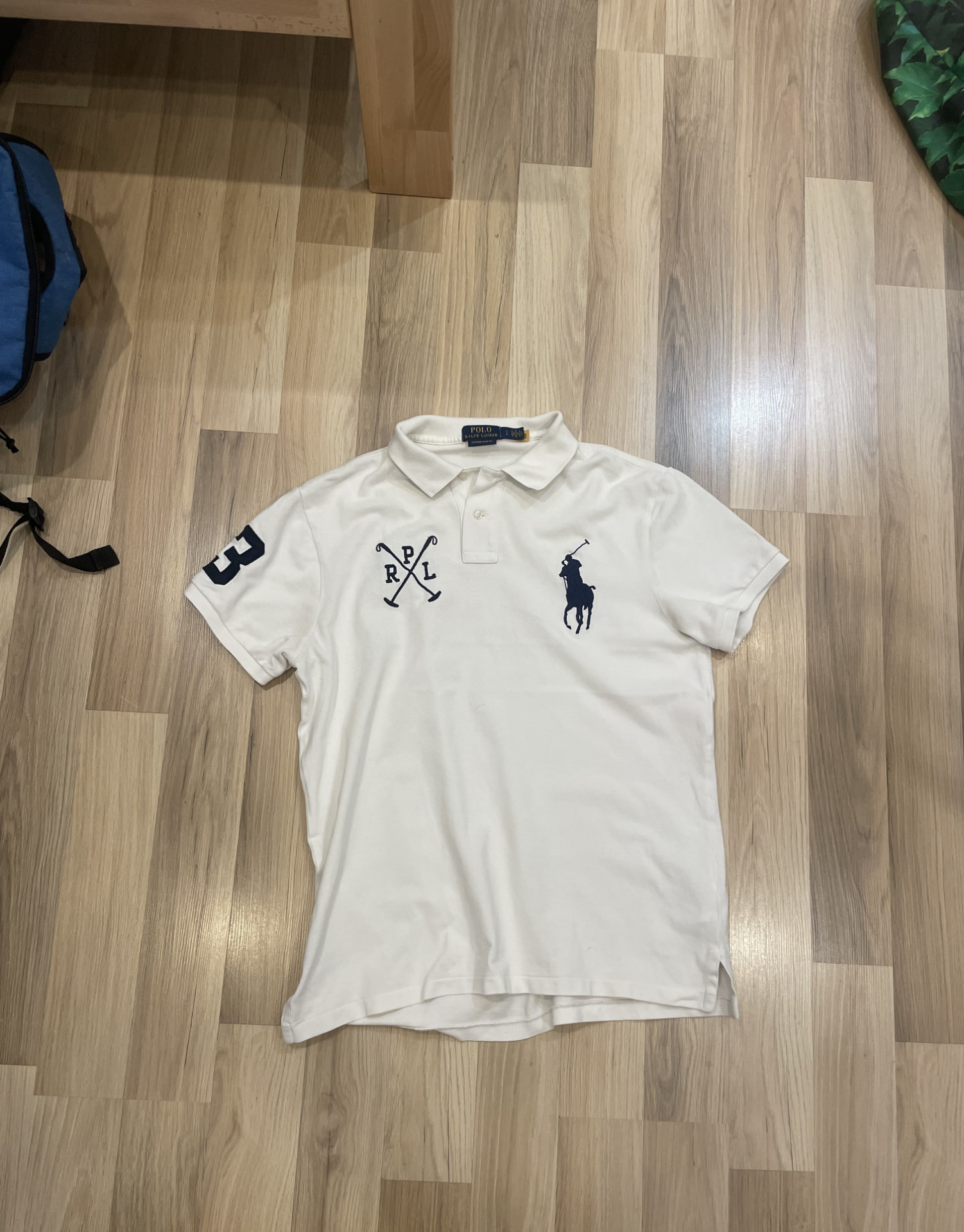 The original phone photo: a white polo shirt lying crooked on a wooden floor with a tote bag beside it under yellow indoor light